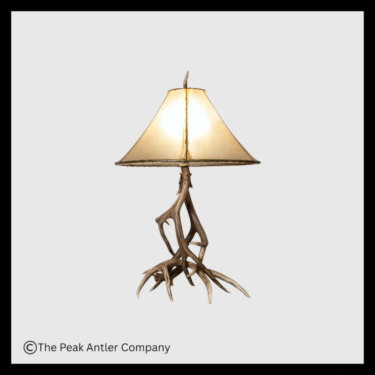 A table lamp with a natural antler base and a warm beige fabric shade, shown against a light gray background with a black border. 