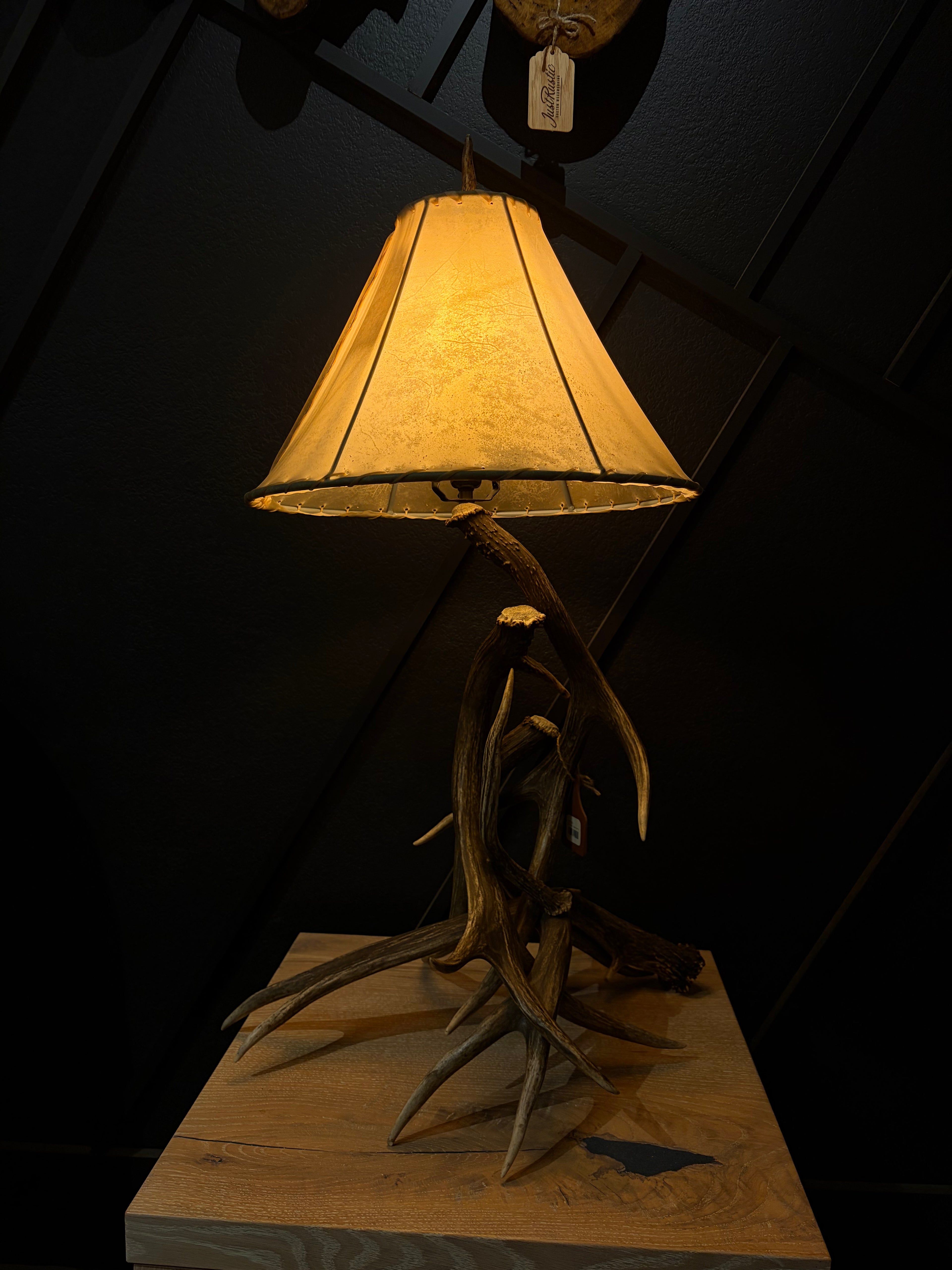 A rustic table lamp with a natural antler base and a warm yellow fabric shade, displayed on a wooden pedestal against a dark paneled background.