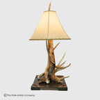 A rustic table lamp featuring a natural antler and driftwood base on a dark rectangular platform, with a cream fabric lampshade, shown against a light gray background.