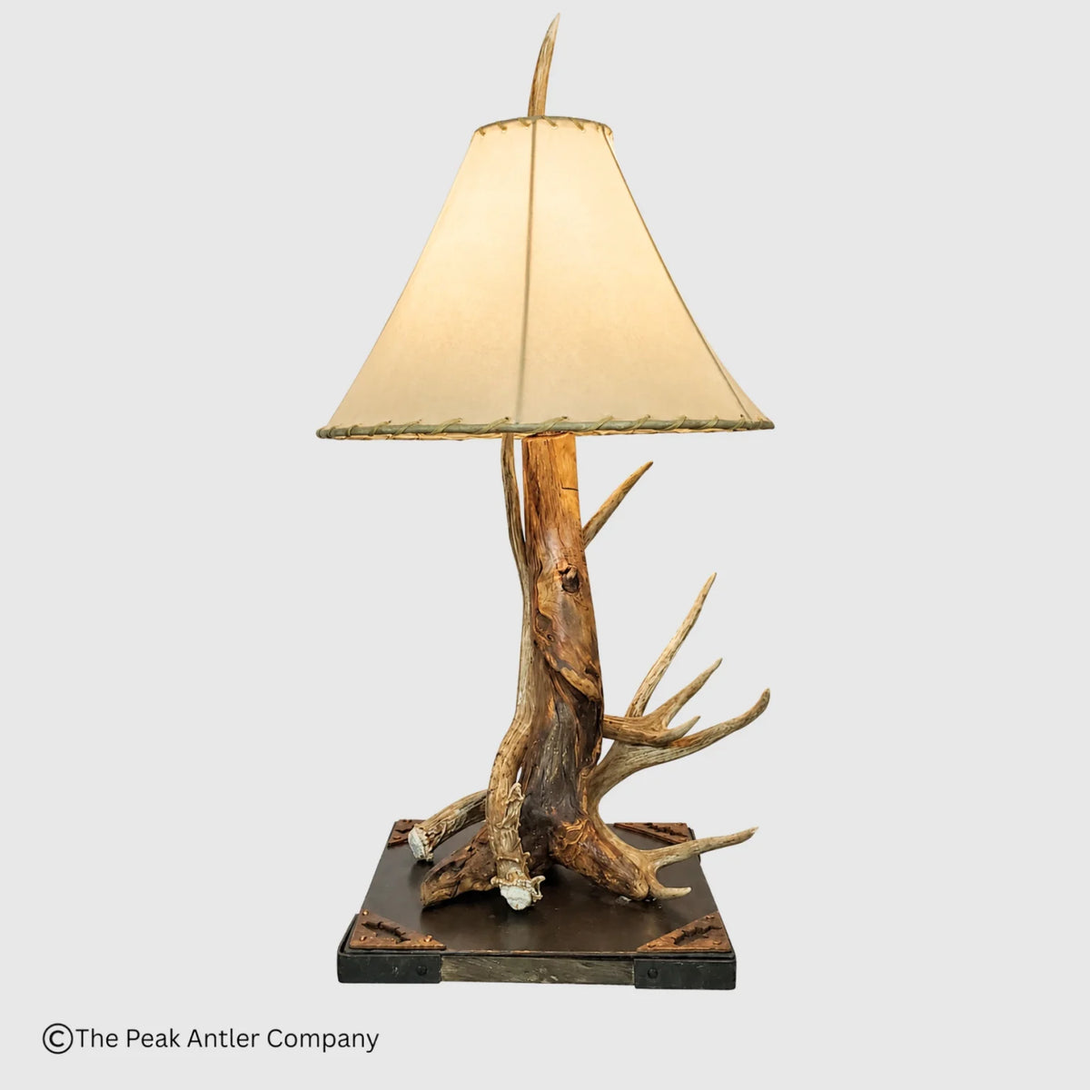 A rustic table lamp featuring a natural antler and driftwood base on a dark rectangular platform, with a cream fabric lampshade, shown against a light gray background.