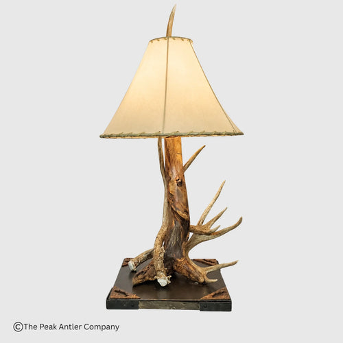 A rustic table lamp featuring a natural antler and driftwood base on a dark rectangular platform, with a cream fabric lampshade, shown against a light gray background.