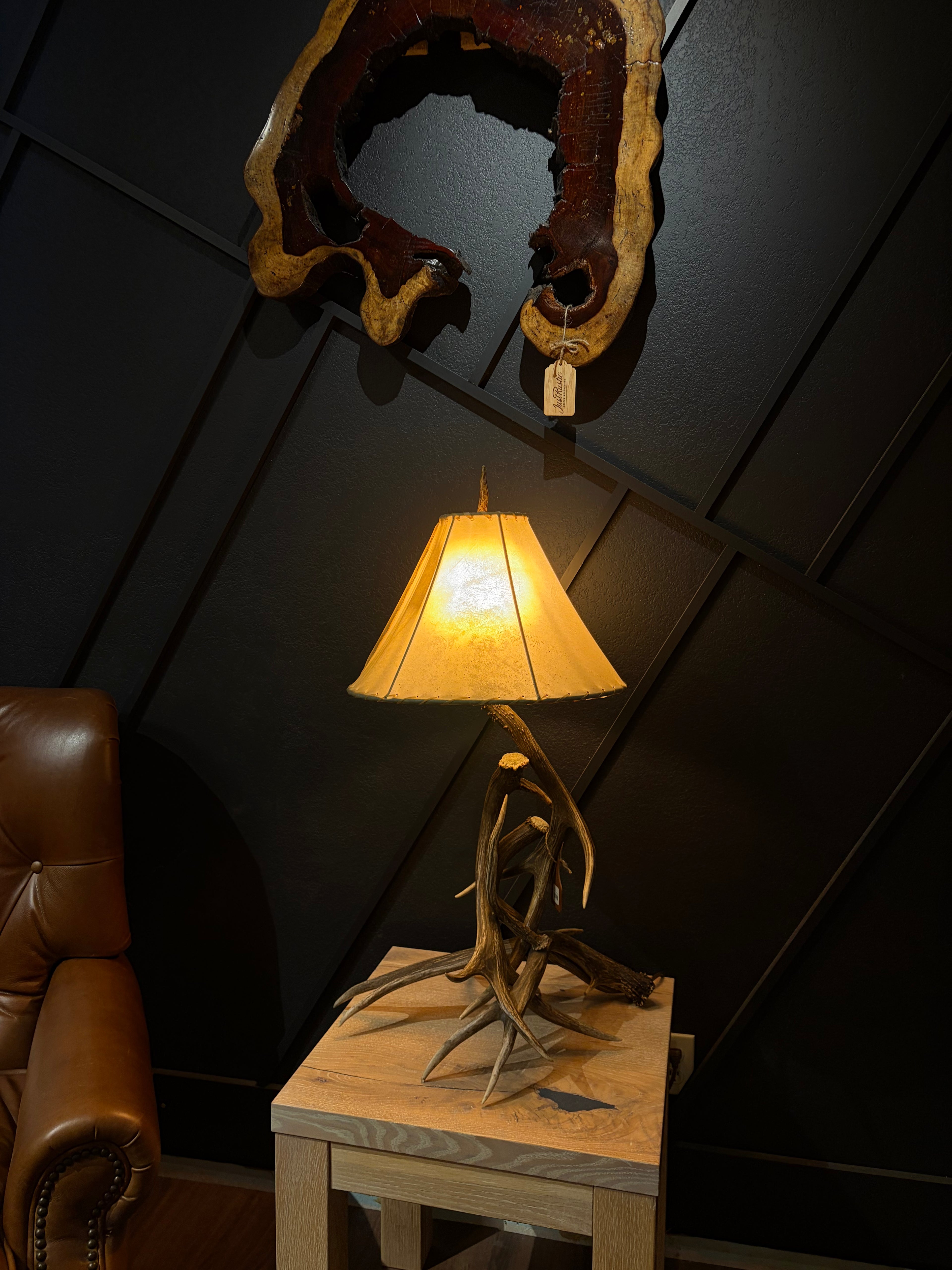 A rustic table lamp with a natural antler base and a warm yellow fabric shade, styled on a light wood side table in a dark room next to a brown leather chair.