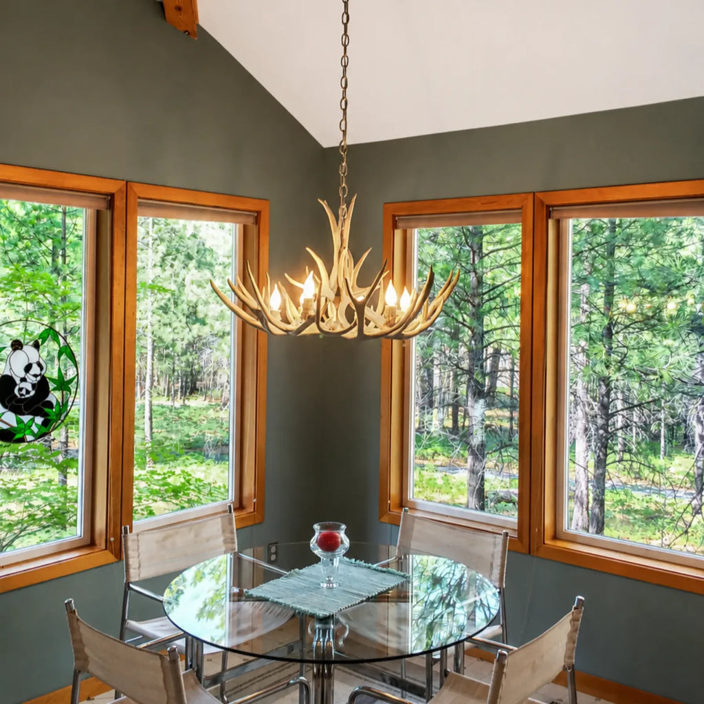 A lighted antler chandelier hangs over a glass dining table in a modern dining room with dark green walls, wood-trimmed windows showing a forest view, and four upholstered chairs.