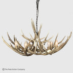 A rustic deer antler chandelier with six lit candle-style bulbs, suspended from a black chain against a light gray background. A copyright watermark for The Peak Antler Company is visible in the bottom-left corner.