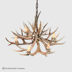 A rustic deer antler chandelier with six lit candle-style bulbs, suspended by a dark metal chain against a light gray background. A copyright watermark for The Peak Antler Company is visible in the bottom-left corner.