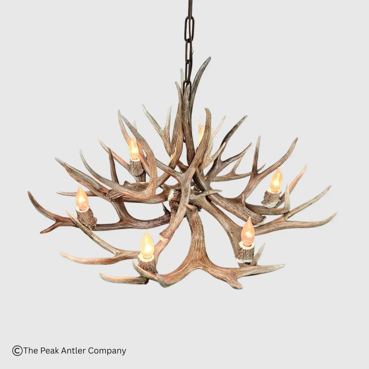 A rustic deer antler chandelier with six lit candle-style bulbs, suspended by a dark metal chain against a light gray background. A copyright watermark for The Peak Antler Company is visible in the bottom-left corner.