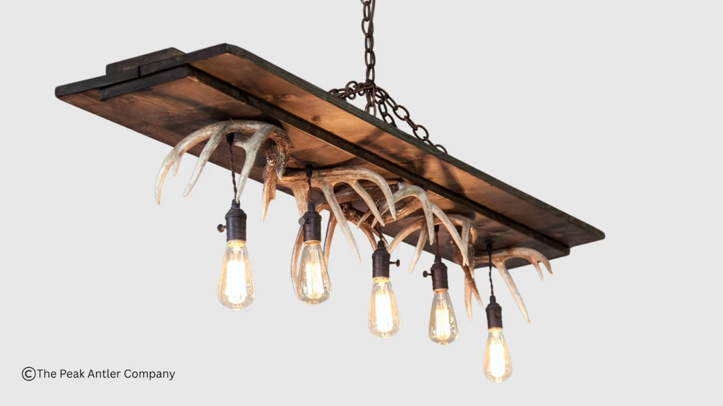 Rustic rectangular wooden chandelier with five hanging antlers and exposed Edison-style bulbs, shown against a light gray background. A small watermark in the lower left reads © The Peak Antler Company.