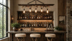 A rustic bar interior with a wooden counter, three upholstered stools, built-in shelving filled with bottles, wall sconces, and a hanging light fixture with antler-style branches and exposed bulbs.