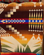 Pendleton blanket with chocolate brown, gold, cream, and green geometric patterns and a blue and red accent stripe.