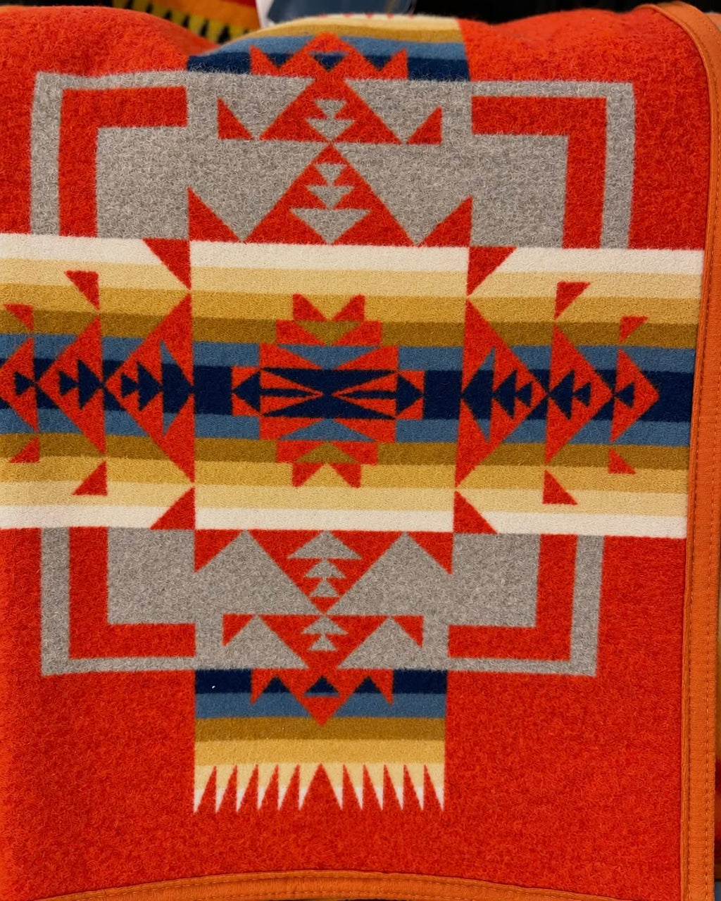 Red-orange Pendleton blanket with blue, navy, gold, cream, and gray Southwestern geometric patterns and orange edging.