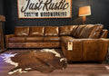 Brown leather sectional sofa in a room with a cowhide rug and 'Just Rustic Custom Woodworking' sign.