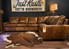 Brown leather sectional sofa in a room with a cowhide rug and 'Just Rustic Custom Woodworking' sign.