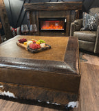 Brown leather ottoman with a tray of food and wine glasses in front of a fireplace.