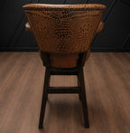 Brown leather barstool with embossed crocodile accents against a black paneled wall.