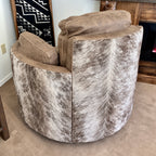 A round swivel recliner with a brindle cowhide-patterned seat and back, and gray leather armrests and back cushions, shown in a home interior setting.